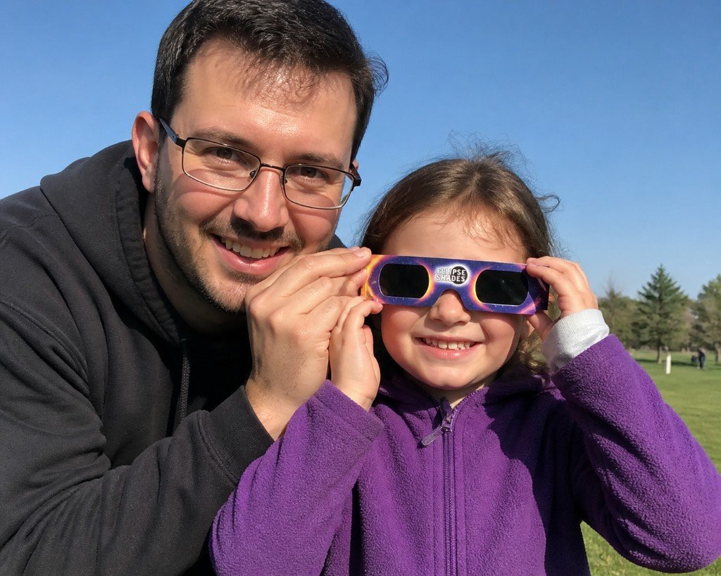 An adult helps a young girl adjust ISO-certified eclipse glasses over her eyes in a sunny field before viewing a solar eclipse.