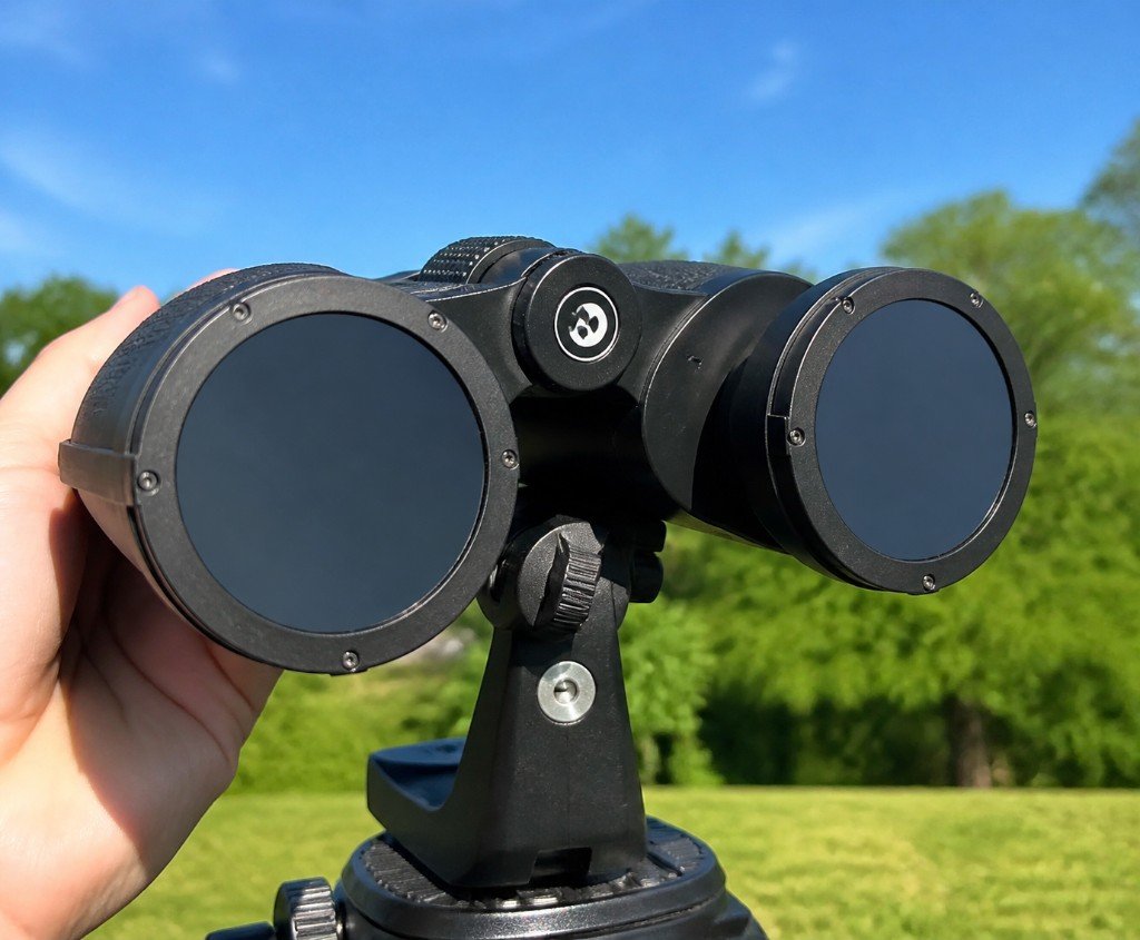 Binoculars on a tripod with dark solar filters fitted over the front objective lenses, held steady in a sunny outdoor field.