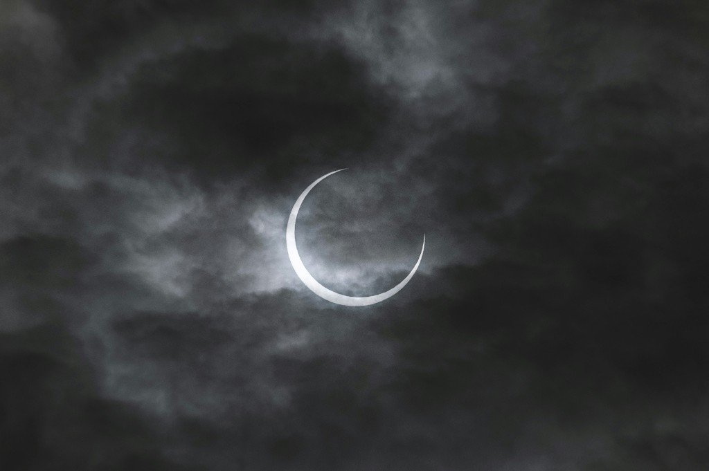 A thin solar crescent barely visible through thick, unmoving grey cloud cover — the eclipse happening unseen above.
