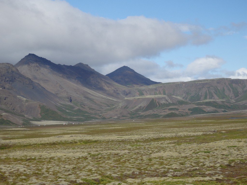 A vast moss-covered plain leading to distant volcanic mountains under a partly cloudy sky.