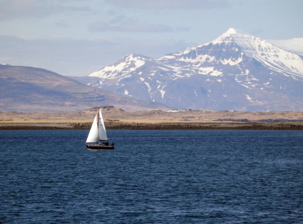 A sailboat on blue water with snow-capped mountains rising beyond the shore.