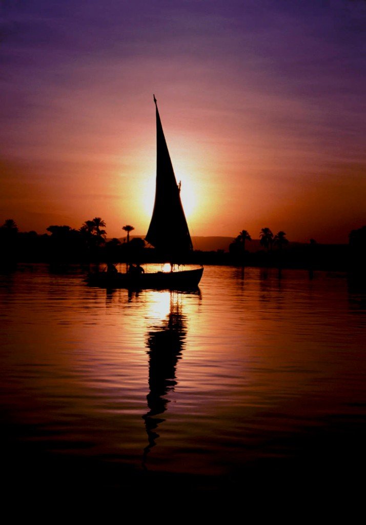 A traditional felucca sailboat silhouetted on the Nile at sunset, with palm trees and hills on the horizon.