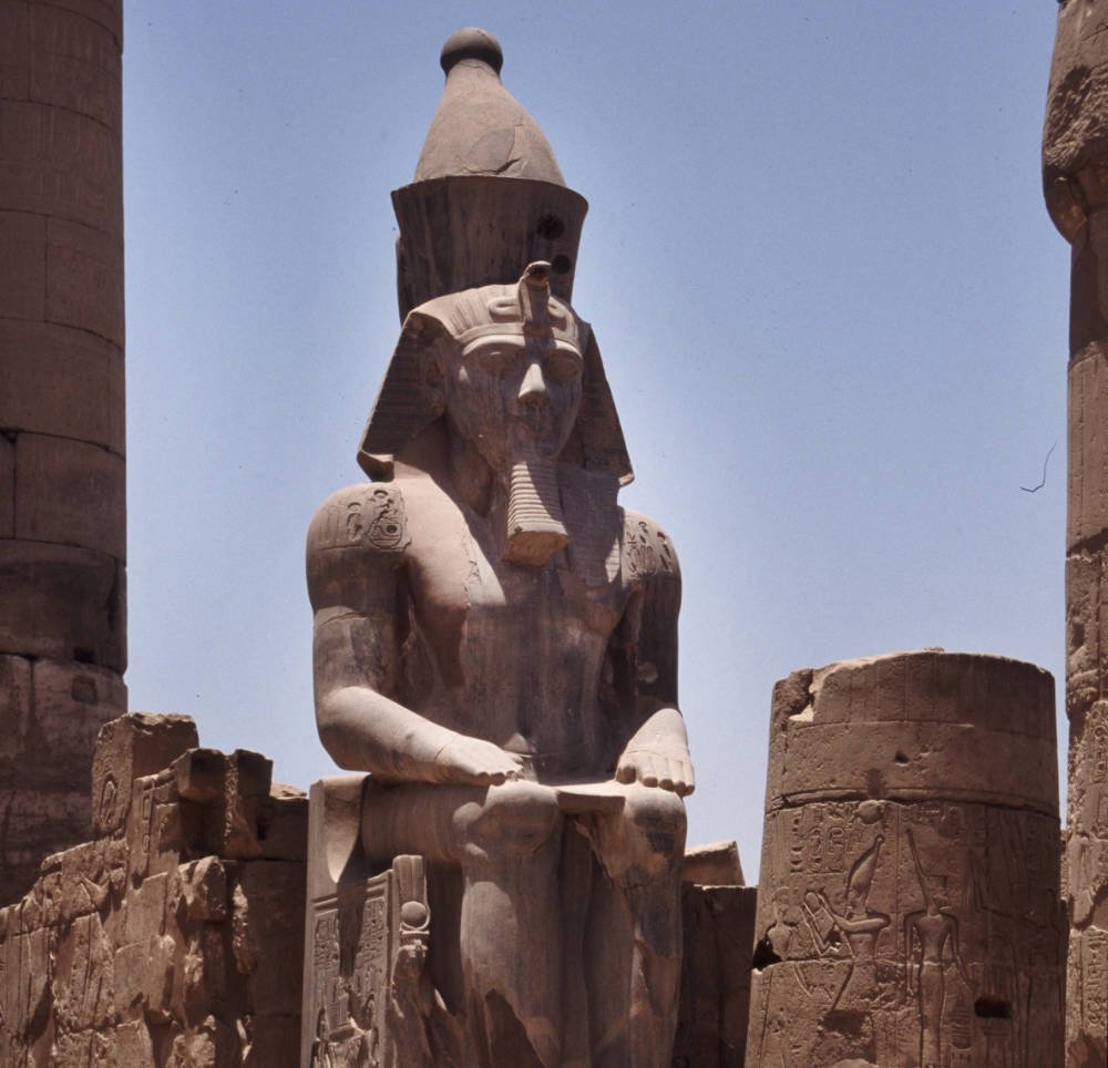 A colossal seated pharaoh statue at Luxor Temple, framed by carved columns and hieroglyphs.