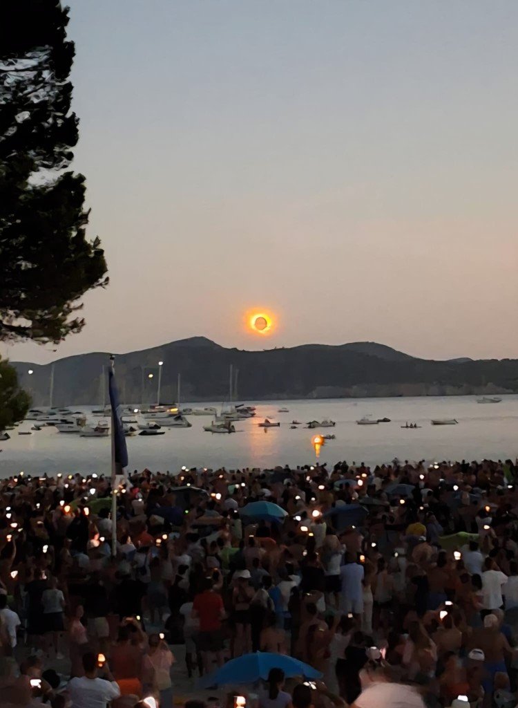 A large crowd on a beach and promenade watches a total solar eclipse at dusk. The black disk of the Moon and bright corona hang low above mountains across a bay, with boats on the water and many observers holding up phones to photograph the sky.