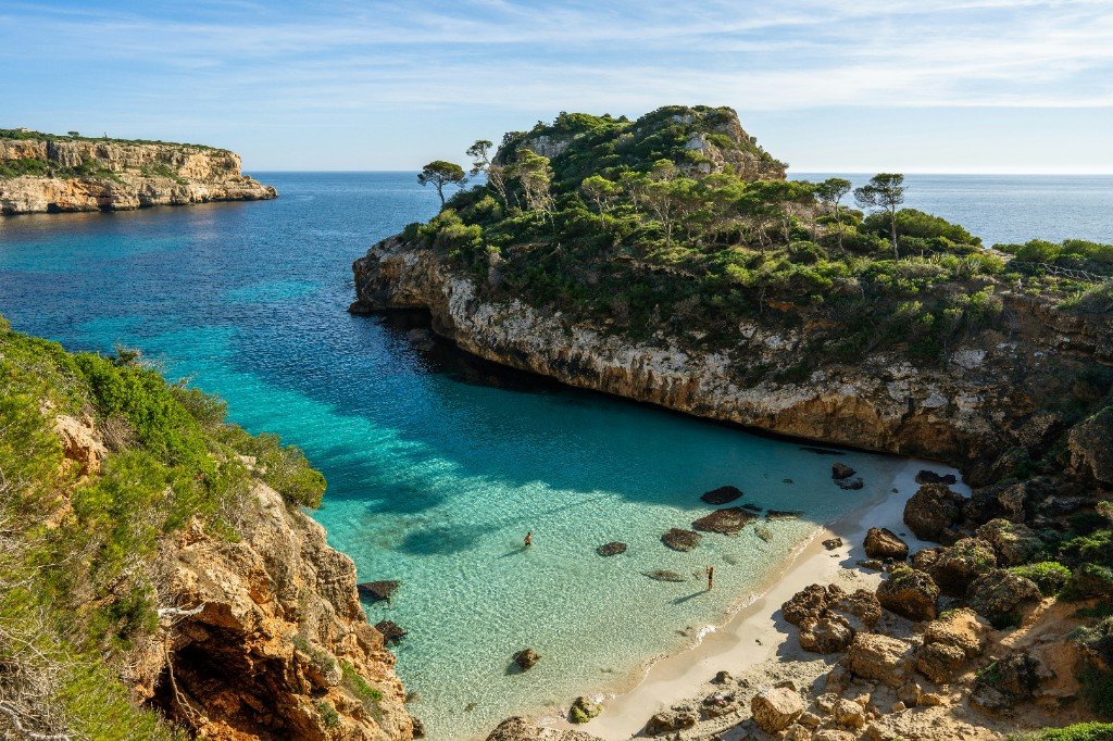 A turquoise Mediterranean cove with sandy beach, pine-covered cliffs, and swimmers in crystal-clear water.