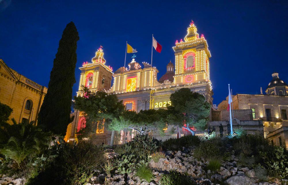 A Maltese parish church in Cospicua lit with festive yellow and red illuminations against the night sky.