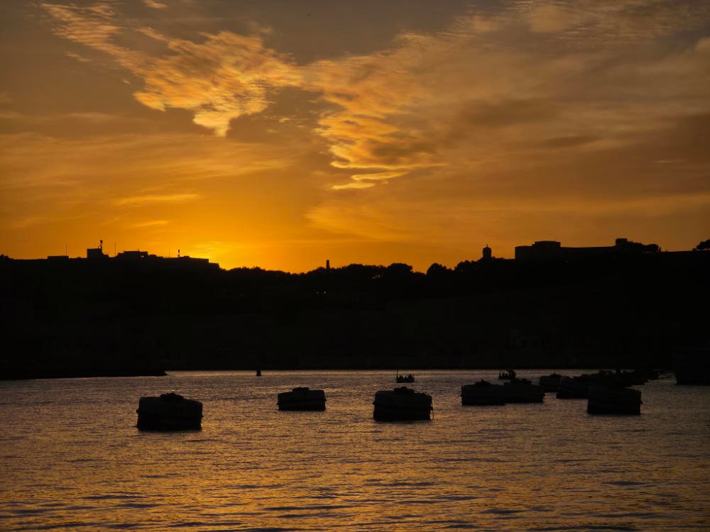 Maltese harbour at sunset, with town silhouettes and orange light reflected on the water.