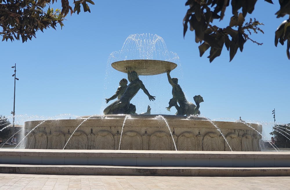 The Triton Fountain at Valletta's City Gate, with bronze figures holding a water basin against a clear blue sky.