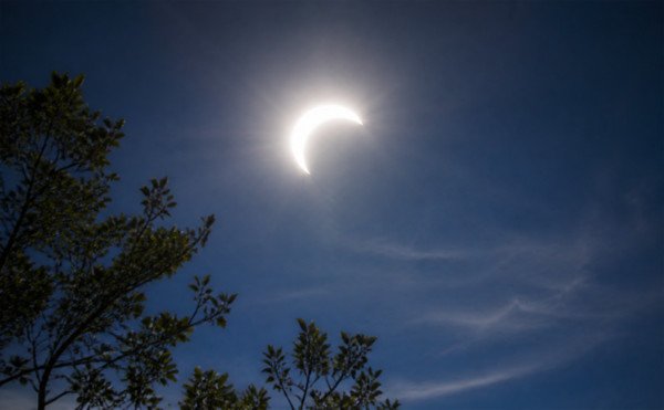Partial solar eclipse: a bright crescent of the Sun still visible in a deep blue sky above silhouetted trees, with intense glare radiating from the exposed limb.